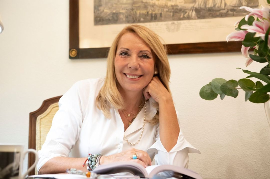 Una mujer rubia con camisa blanca sonríe mientras está sentada en un escritorio