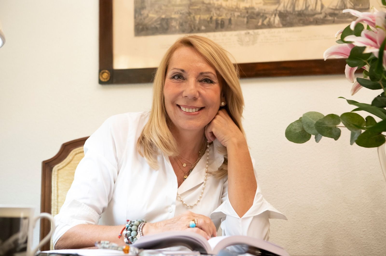 Una mujer rubia con camisa blanca sonríe mientras está sentada en un escritorio