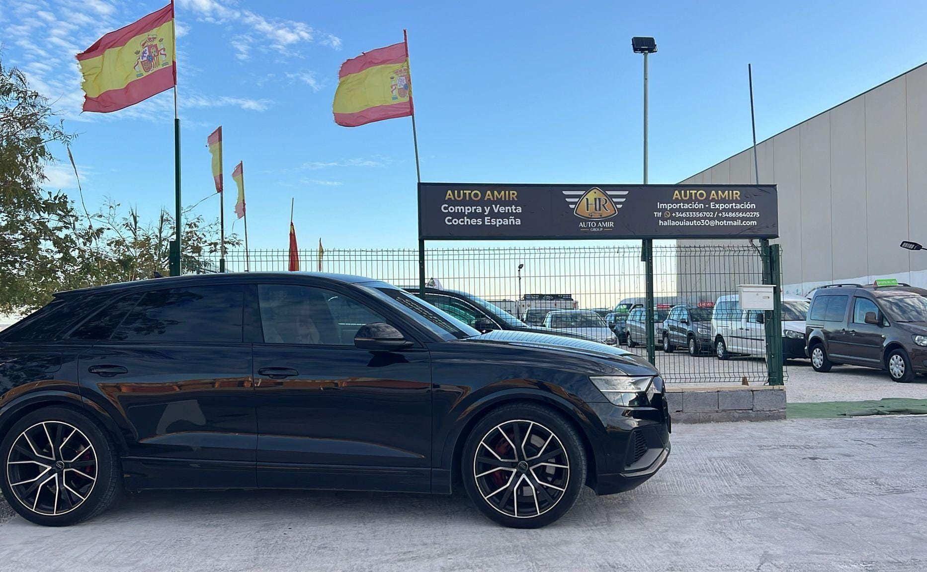 Un SUV negro estacionado frente a un concesionario de automóviles con banderas españolas ondeando al fondo.