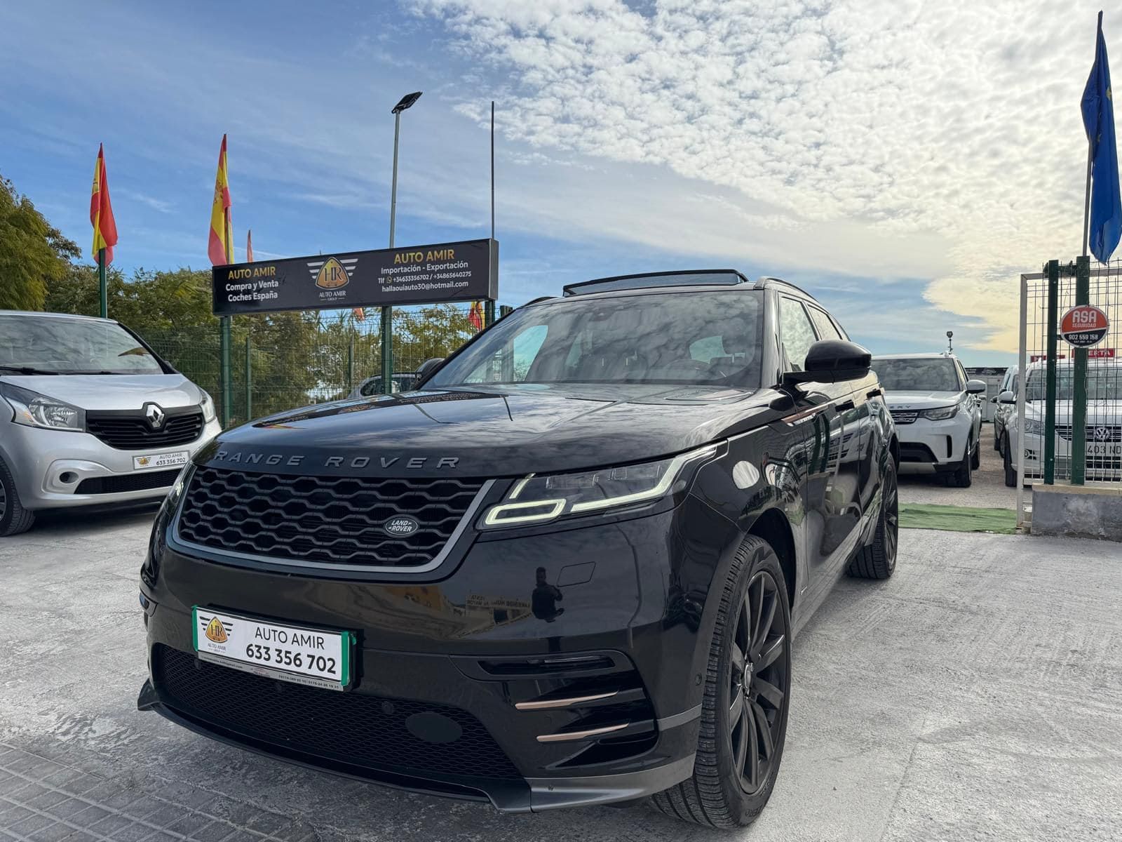 Un Range Rover negro estacionado en el aparcamiento de un concesionario de coches, con banderas y otros vehículos al fondo.