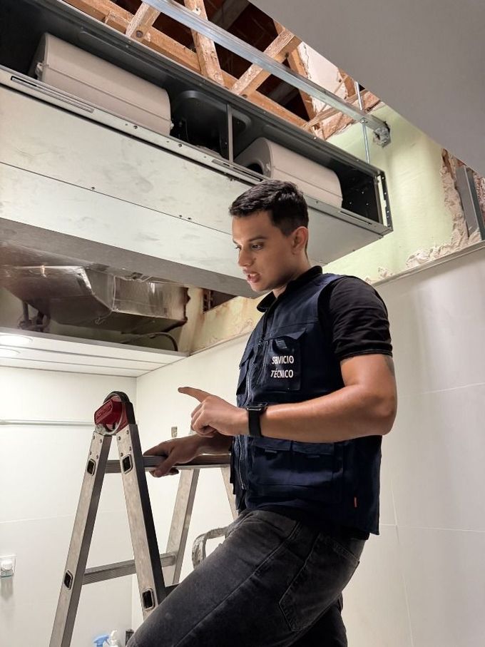 Un hombre utiliza una escalera para inspeccionar los conductos de ventilación del techo en un cuarto de servicio estrecho.