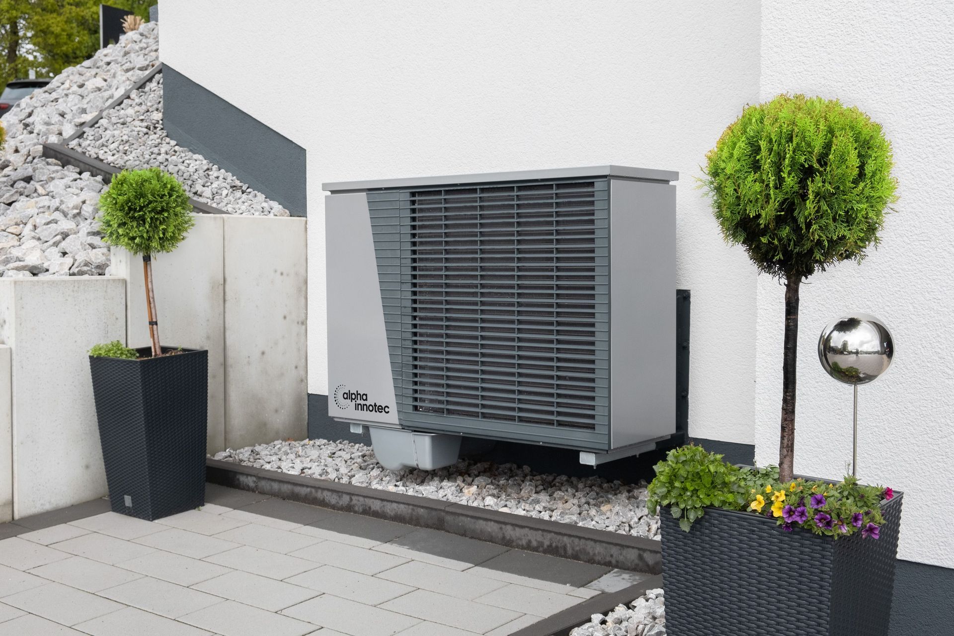 Unidad de aire acondicionado exterior junto a una pared blanca, con plantas en macetas y grava en un patio moderno.
