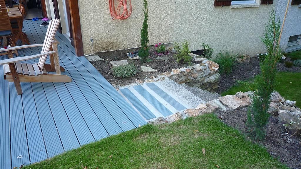 Terrasse grise avec escalier menant à une cour gazonnée jouxtant un mur texturé orné de plantes et d'un petit arbre.