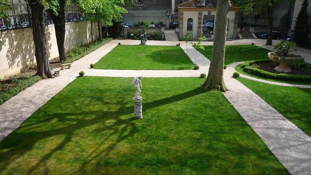 Jardin à la française avec pelouse verte, allées de pierre, arbres et un petit bâtiment en arrière-plan.