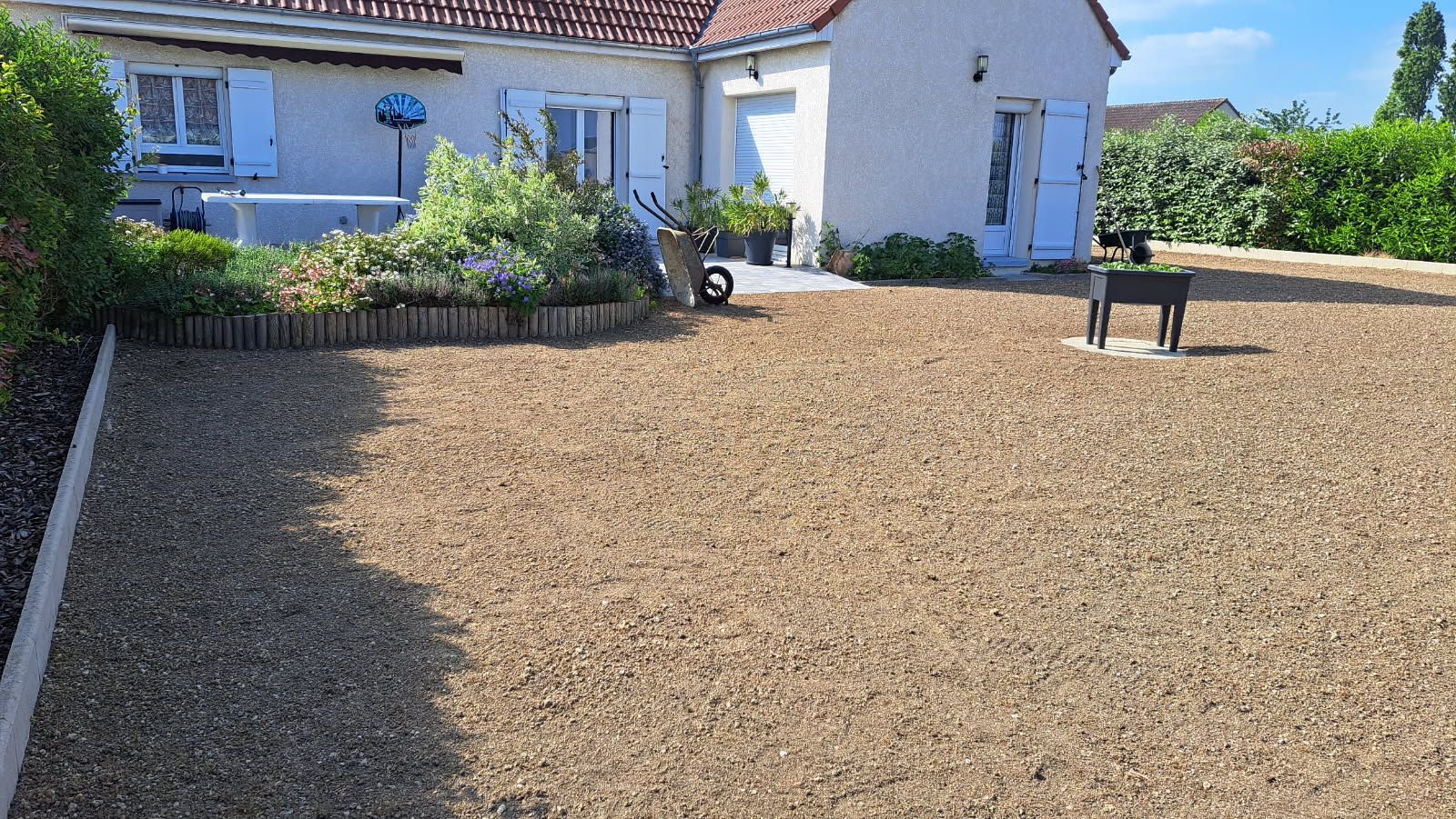 Cour de gravier devant une maison claire avec un petit parterre de fleurs et un barbecue. Ciel bleu.