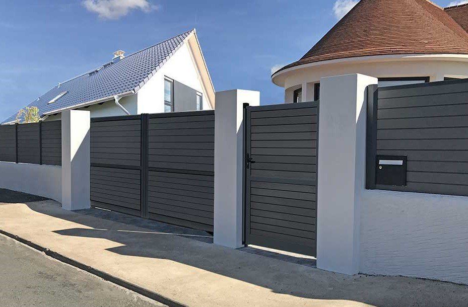 Clôture et portail à lames horizontales grises devant les maisons ; piliers et ornements blancs, ciel bleu.