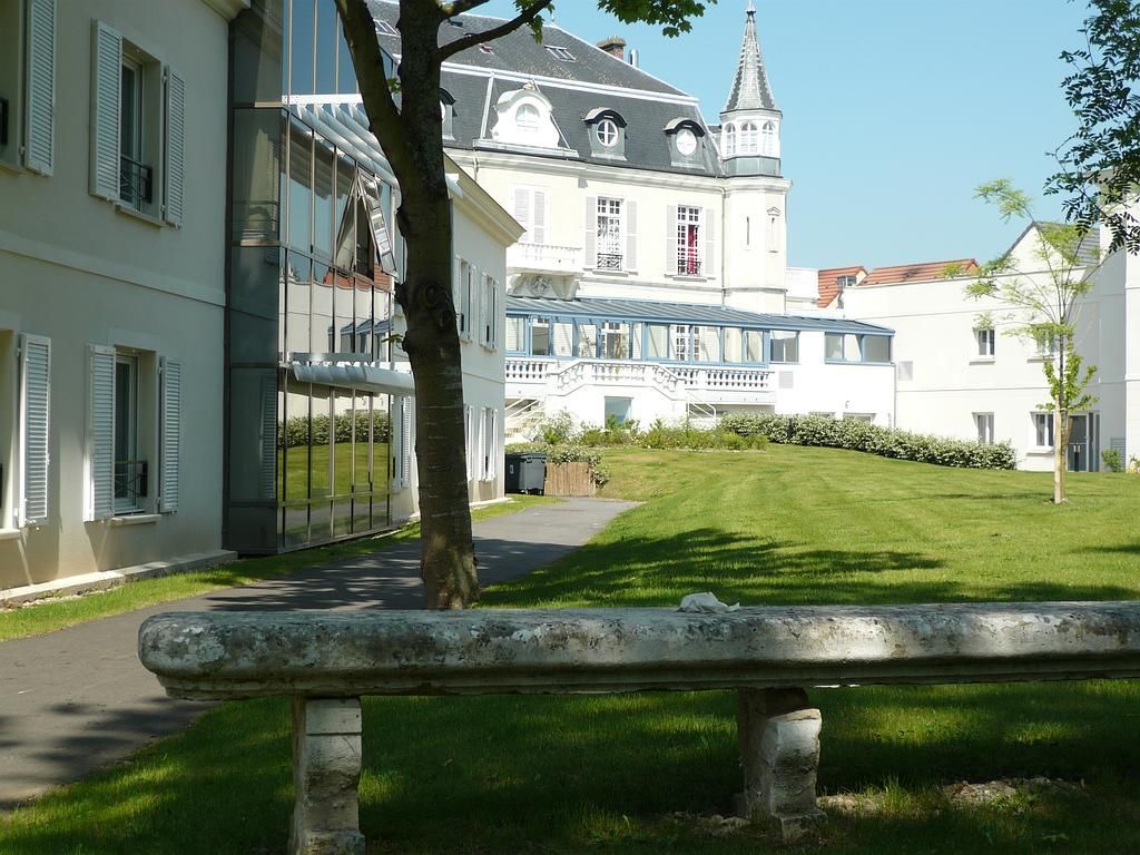 Vue extérieure de bâtiments blancs, d'une pelouse et d'un banc en pierre par une journée ensoleillée. Un arbre se trouve au centre.