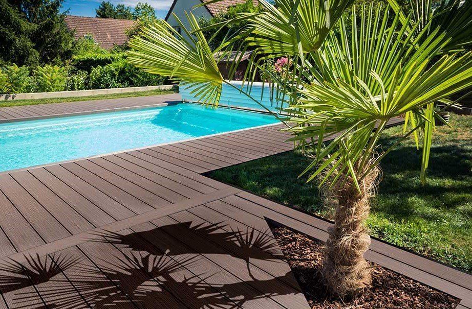 Vue sur la piscine avec terrasse en bois brun, eau turquoise, palmier et pelouse.