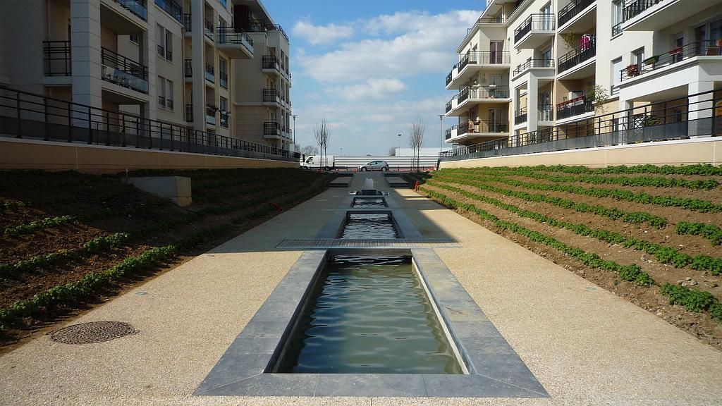 Aménagement aquatique entre les immeubles. Des bassins rectangulaires dévalent un chemin en pente bordé de végétation.