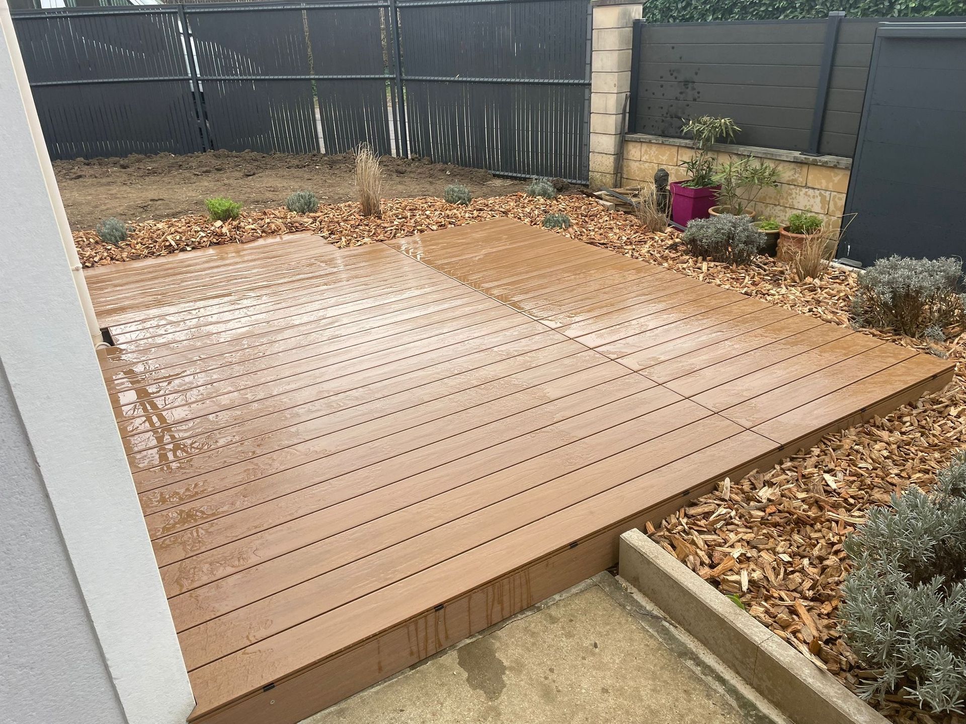 Une terrasse en bois composite brun clair se trouve dans une cour arrière, à côté d'un chemin en béton, entourée de paillis de copeaux de bois et d'une clôture.