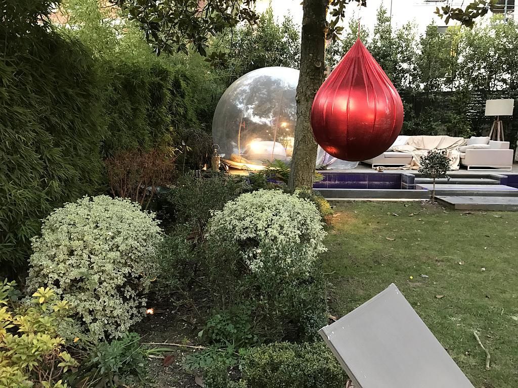 Un ballon rouge en forme de larme est suspendu à un arbre. Une sphère miroir et des arbustes verts se dressent en arrière-plan.