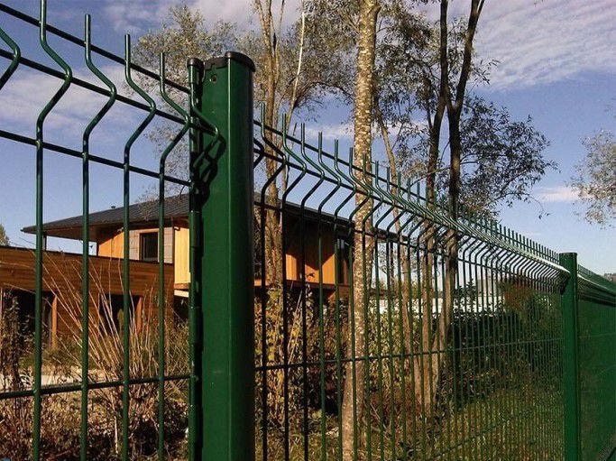 Clôture en fil de fer vert entourant une maison et des arbres par une journée ensoleillée.