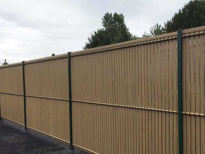 Clôture à lattes verticales beiges avec des poteaux verts, longeant une surface pavée, sous un ciel nuageux.