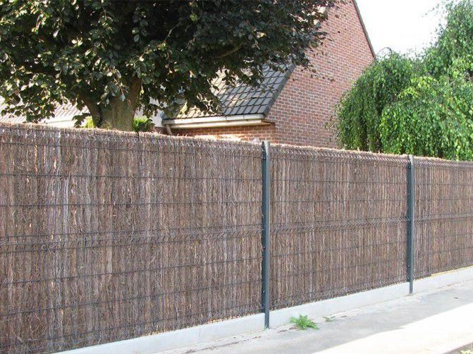 Clôture en roseaux le long d'un trottoir, avec des poteaux en métal sombre et une maison en briques en arrière-plan.