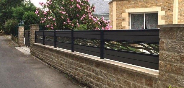 Une clôture noire ornée de découpes décoratives surplombe un mur de pierre le long d'une route, avec de la verdure et un bâtiment en arrière-plan.