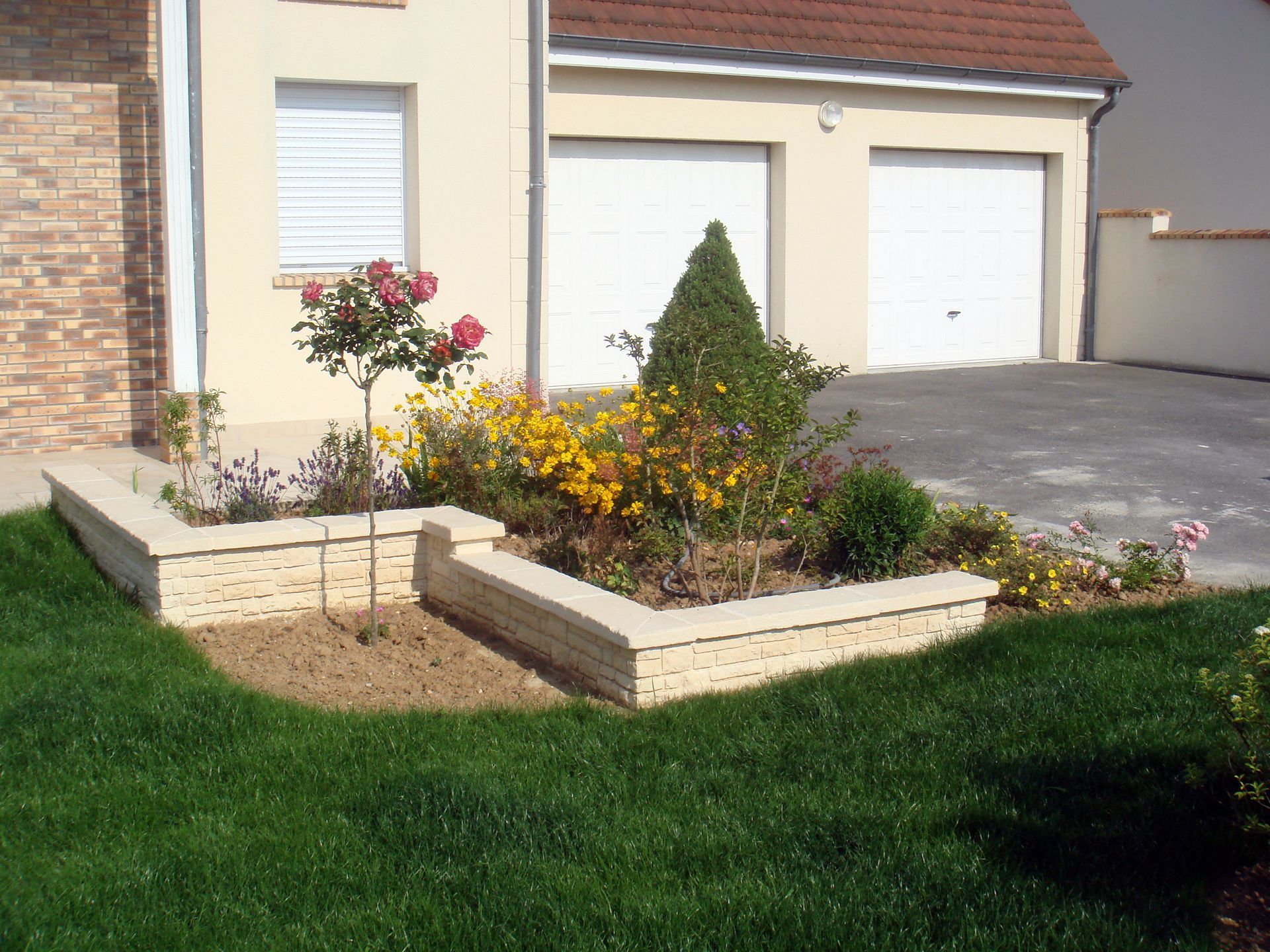 Des parterres de fleurs surélevés, agrémentés de verdure, devant une maison aux portes de garage blanches.