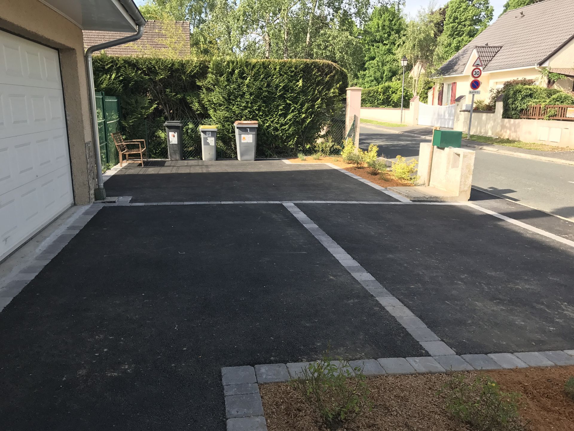 Allée en asphalte noir bordée de gris, poubelles et boîte aux lettres en bord de route, près d'une rue.