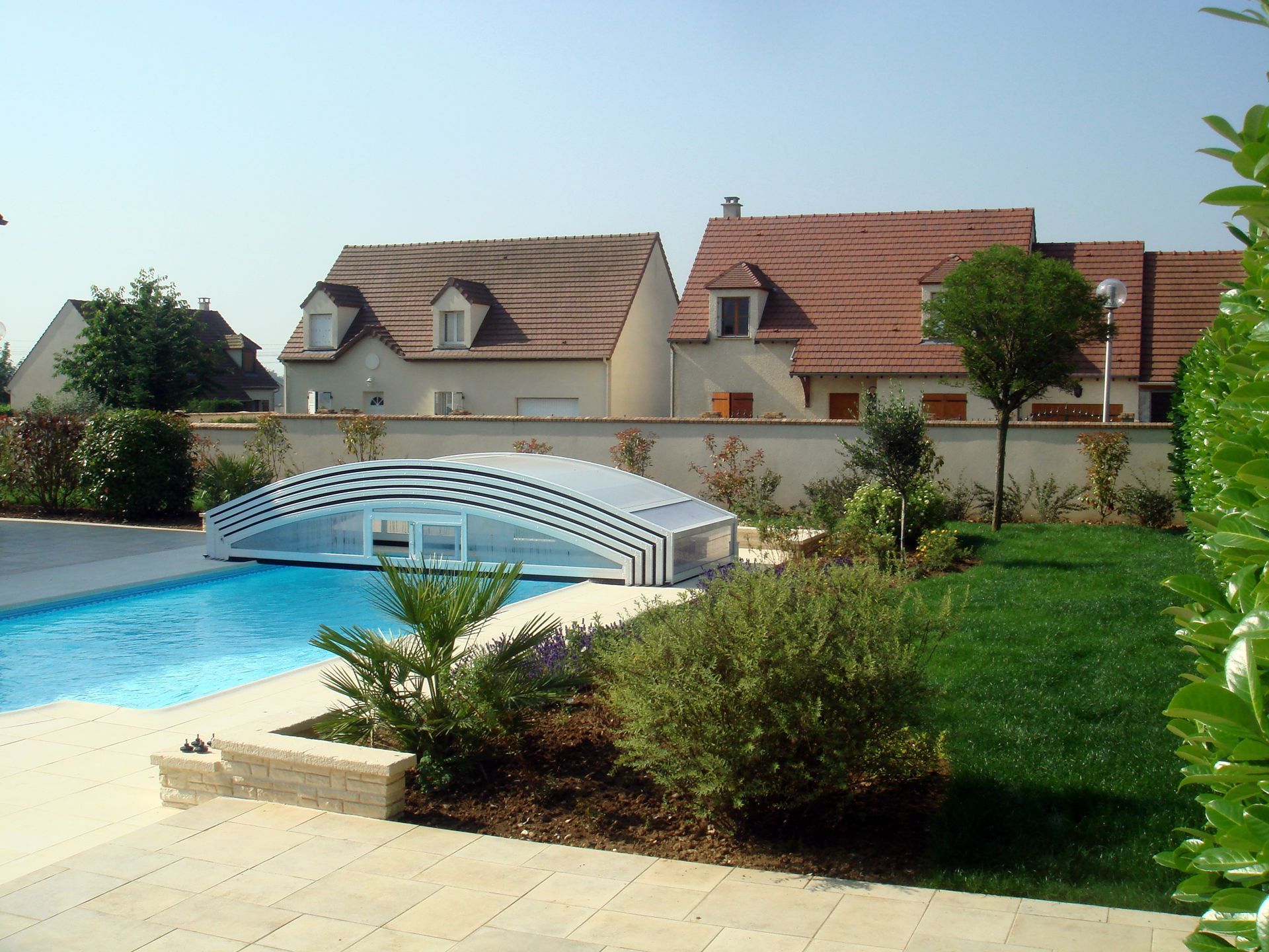 Piscine avec couverture rétractable en forme d'arche dans un jardin, maisons en arrière-plan, journée ensoleillée.