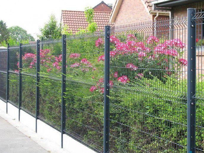 Clôture en fil de fer noir avec des fleurs roses et des buissons verts devant une maison en briques.