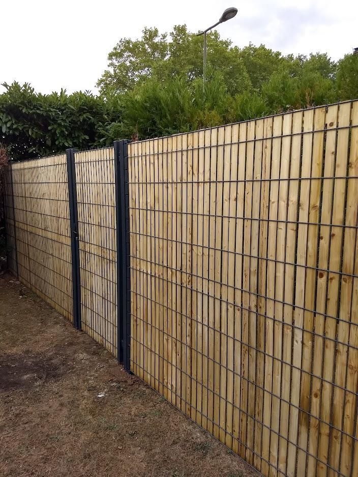 Clôture en bois avec poteaux métalliques, lattes verticales et motif en grille dans une cour avec des arbres en arrière-plan.