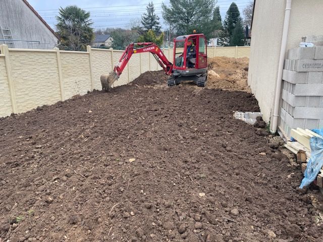 Une mini-pelle creuse dans un terrain en terre battue, à côté d'une clôture et d'un bâtiment.