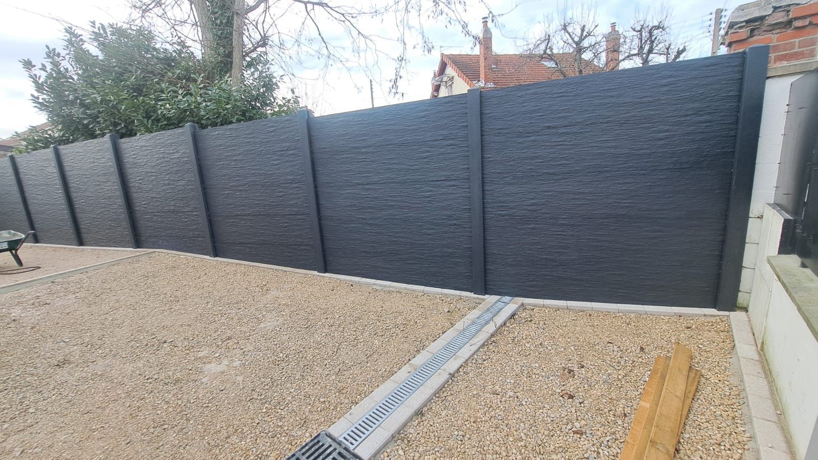 Clôture en béton texturé noir dans une cour en gravier avec un canal de drainage.