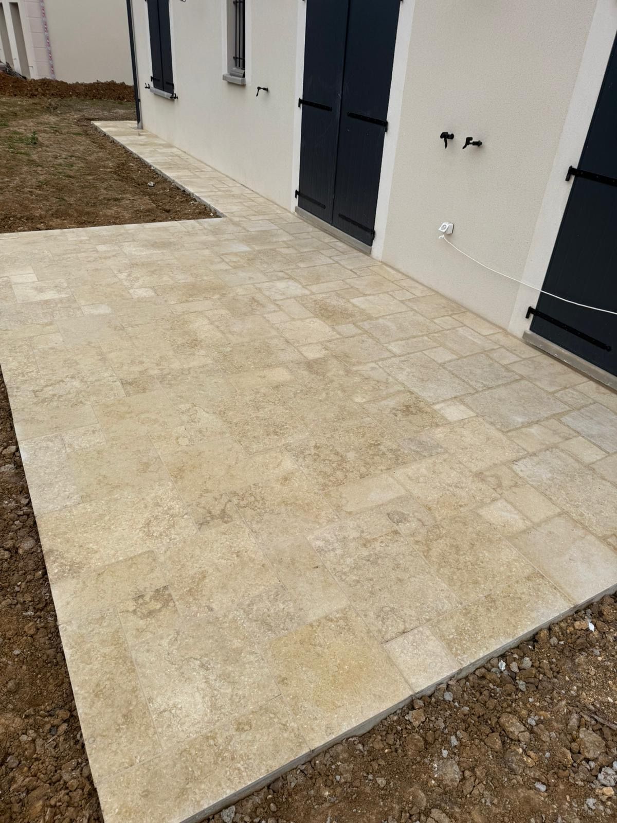 Terrasse en pierre jouxtant un bâtiment couleur crème aux volets foncés.