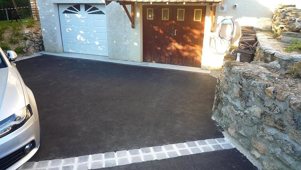 Allée avec une voiture argentée garée à gauche, deux portes de garage et un mur de pierre à droite.