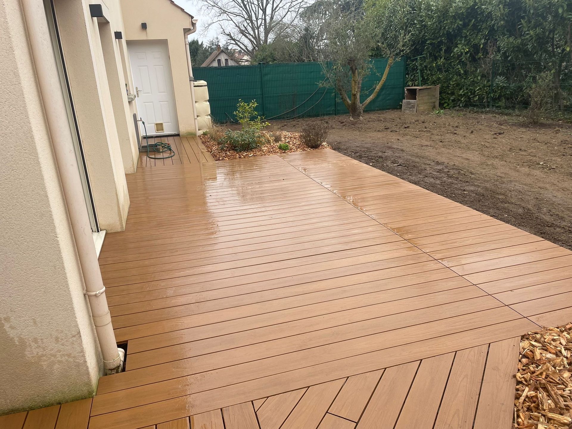 Une terrasse en bois composite brun clair s'étendant d'une maison à une cour bordée d'une clôture verte assurant l'intimité.