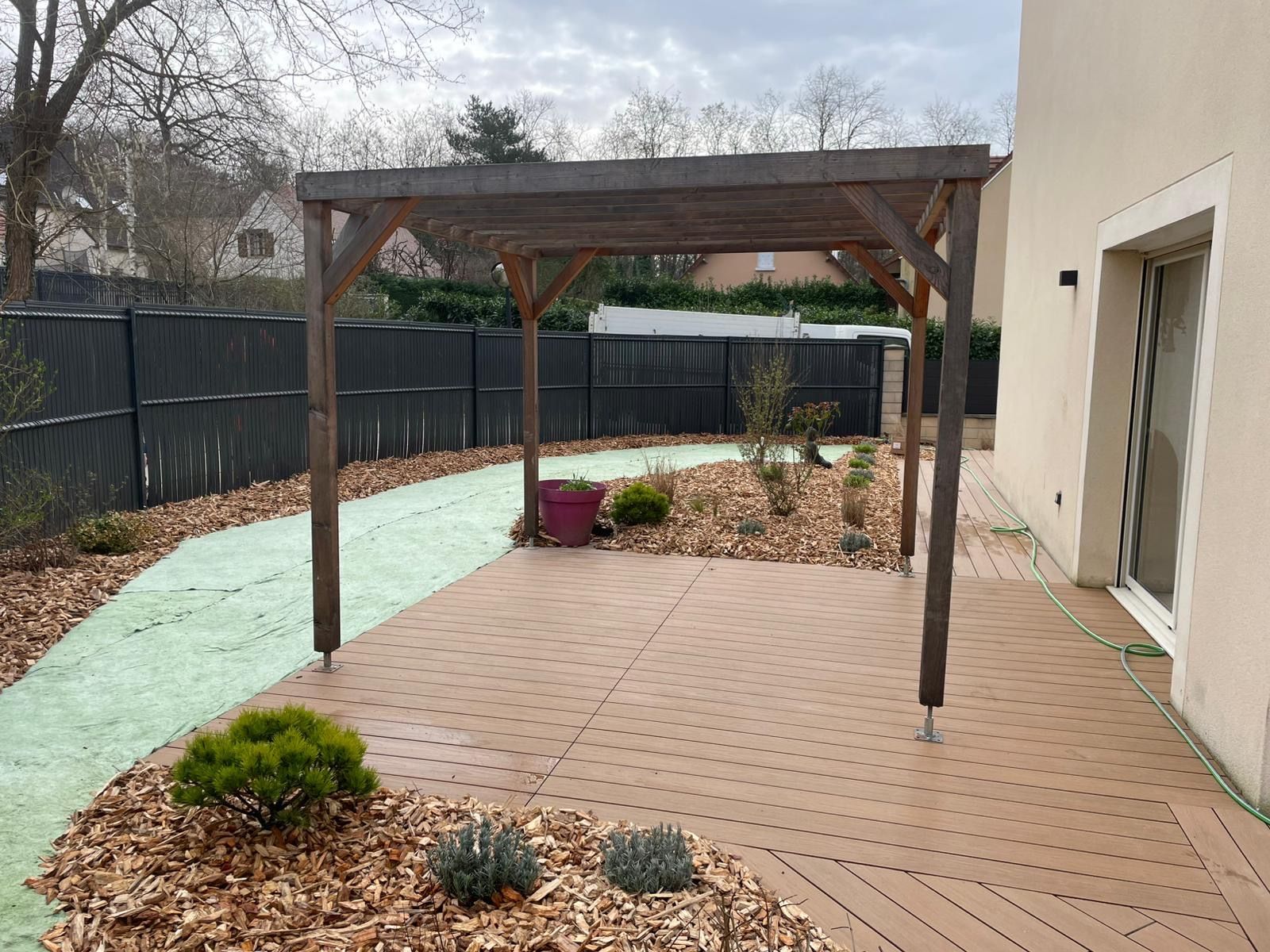 Une pergola en bois brun se dresse sur une terrasse beige clair, à côté d'une maison beige, avec un chemin sinueux et des parterres de fleurs dans la cour.