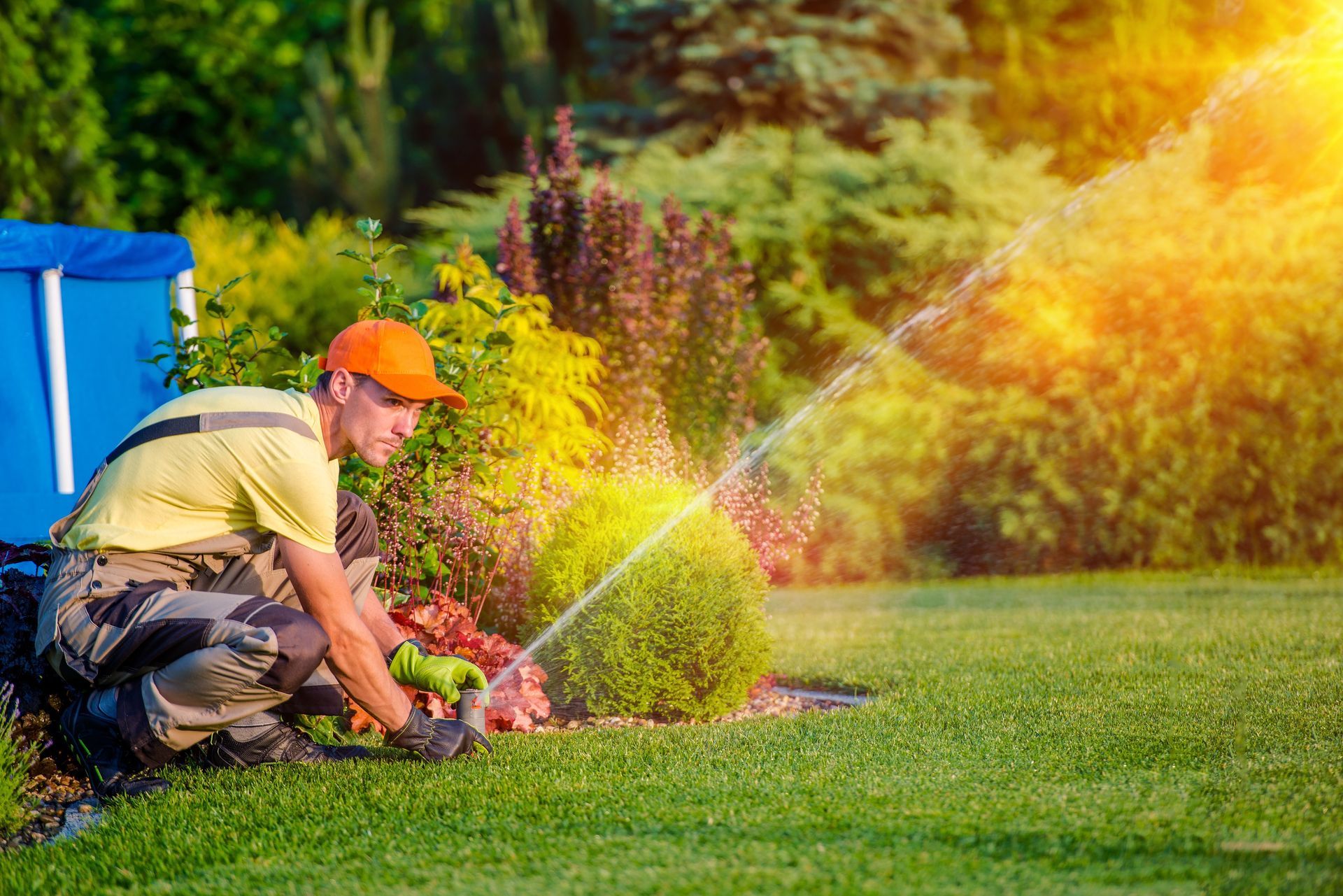 Un homme portant une casquette orange arrose des plantes avec un tuyau d'arrosage dans un jardin ensoleillé.