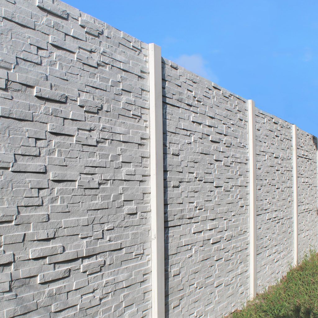 Une haute clôture en béton gris clair, ornée d'un motif de pierres empilées texturées, se détache sur un ciel d'un bleu éclatant.