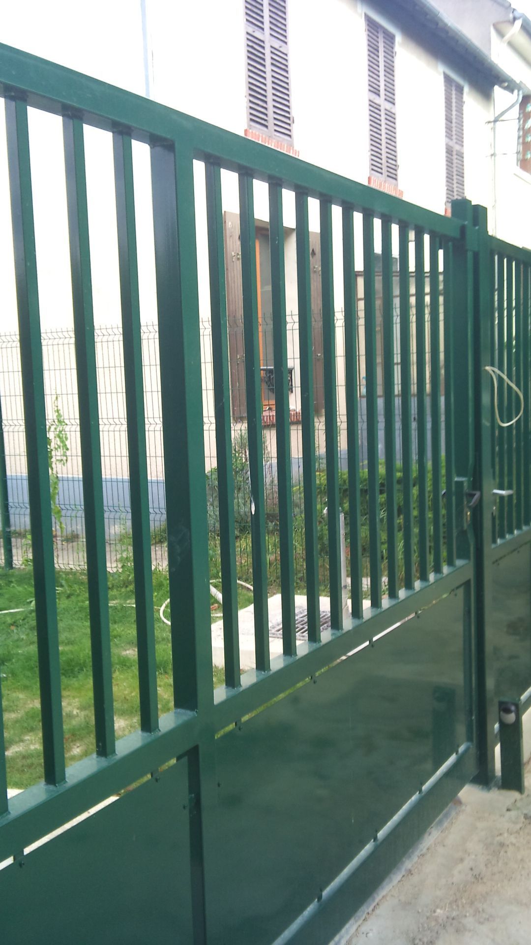 Portail métallique vert à barreaux verticaux, partiellement ouvert, devant un bâtiment.
