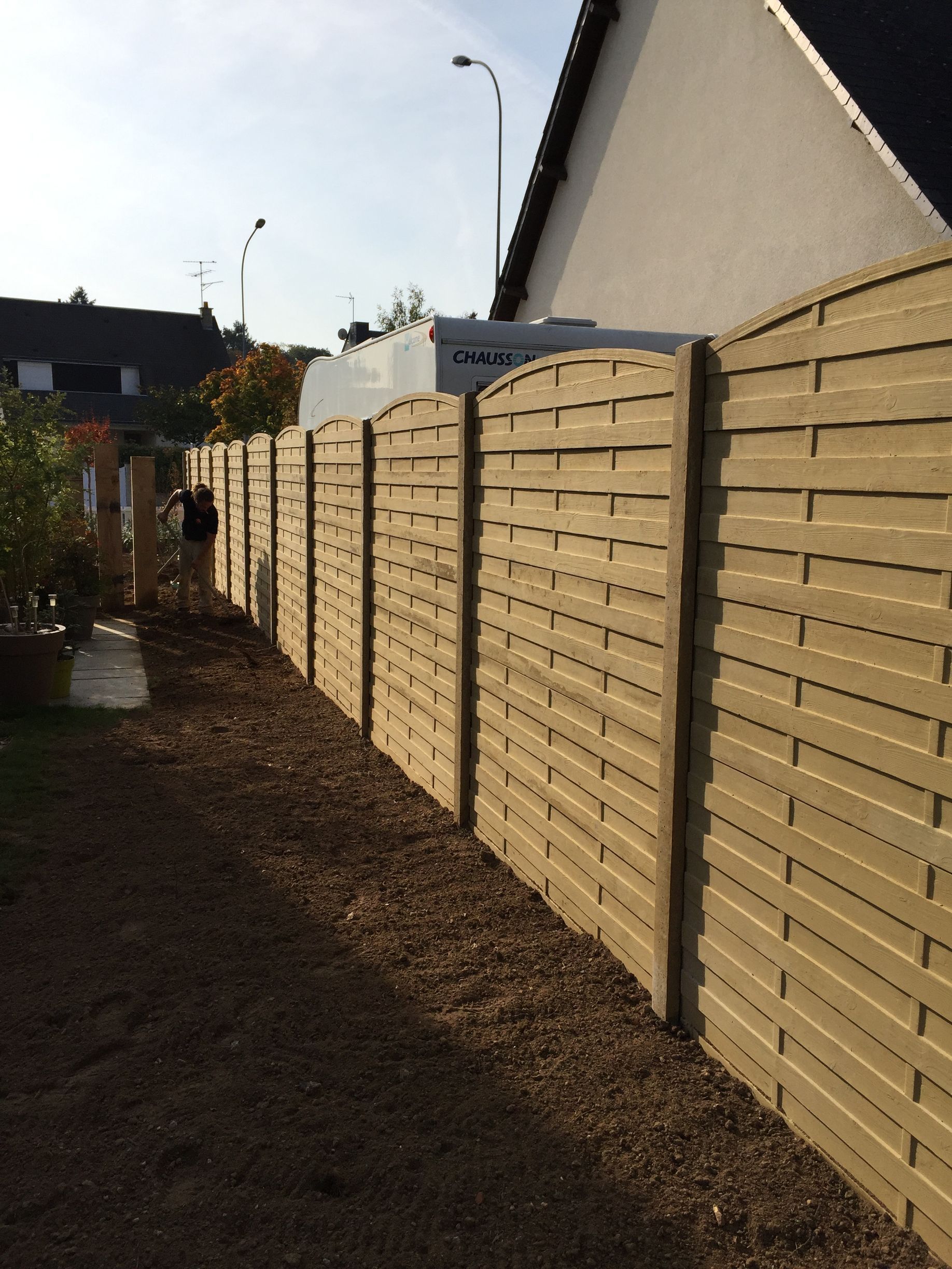 Clôture en bois dans une cour, chemin de gravier à côté. Personne au loin.
