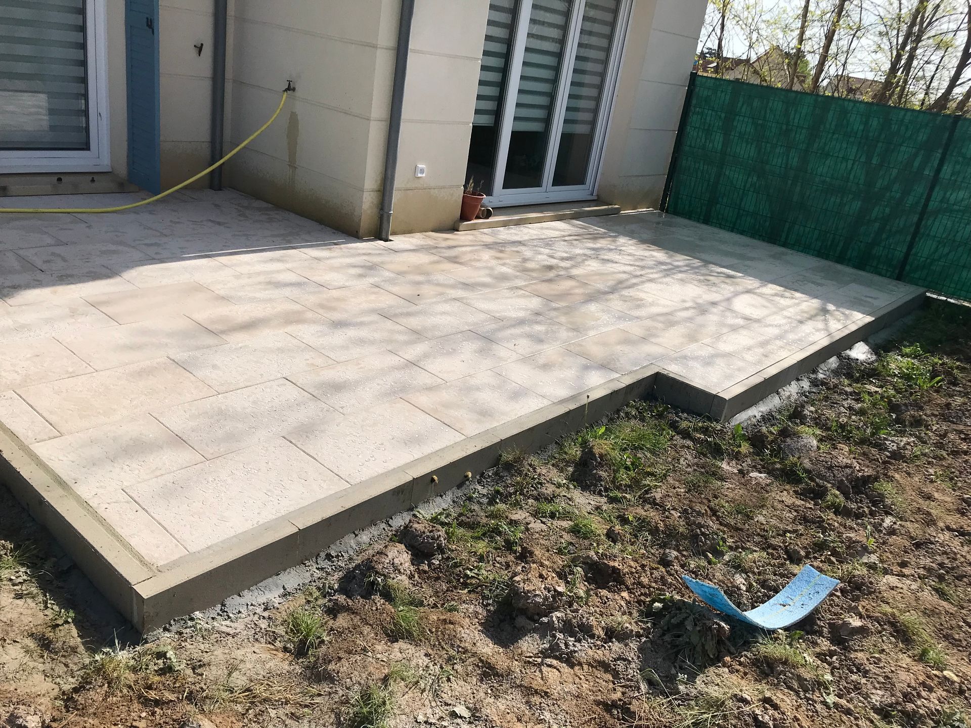 Une terrasse pavée jouxte une maison avec une bordure en béton, adjacente à une clôture verte et à une zone gazonnée.
