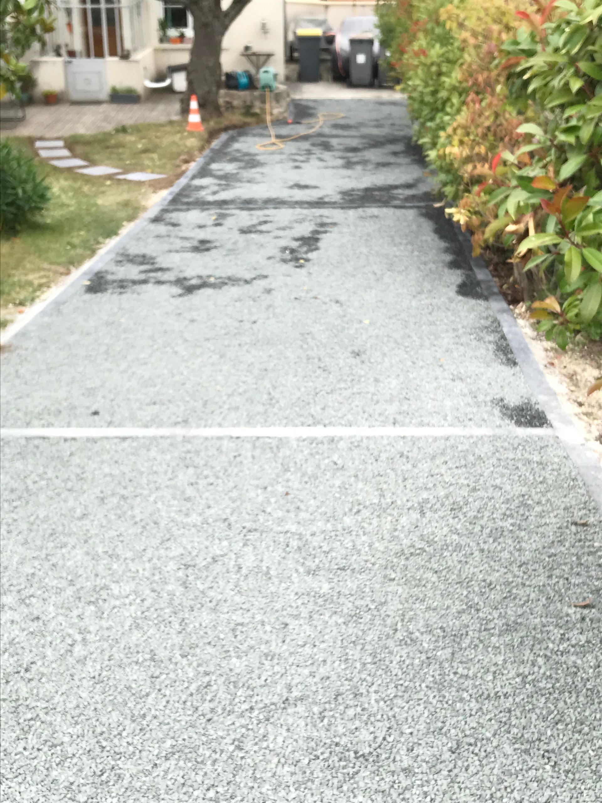 Allée de gravier menant à une maison, bordée de gazon et d'arbustes. Tons gris et verts.