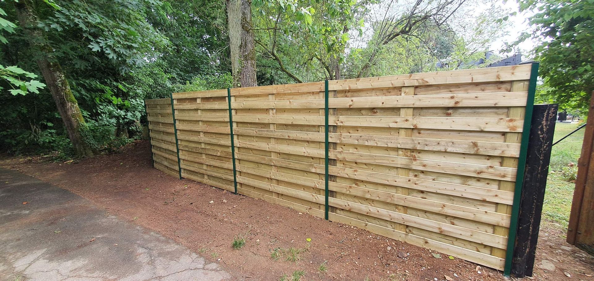 Une clôture en bois avec des poteaux verts longe un chemin de gravier bordé d'arbres.