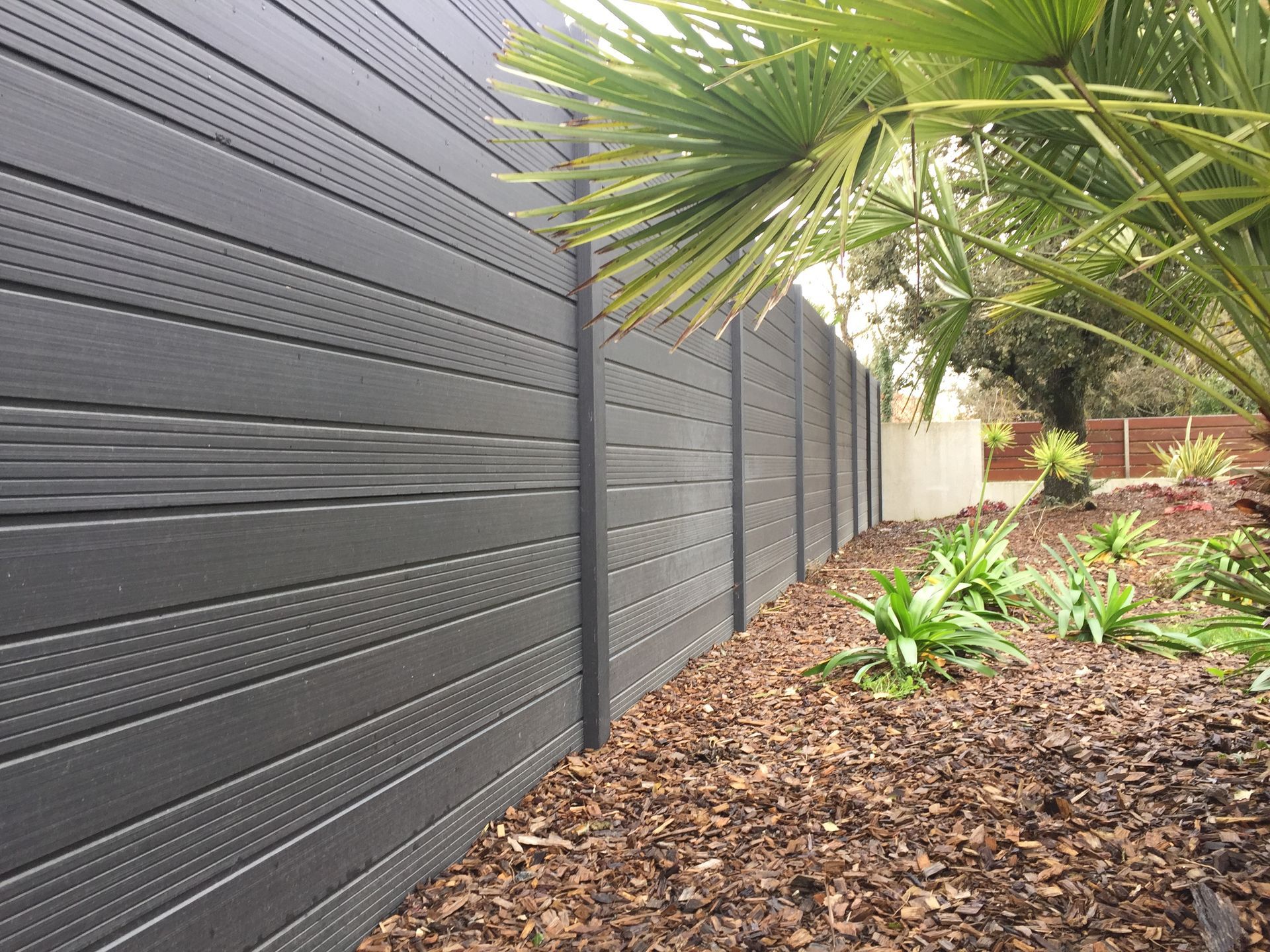 Une haute clôture composite gris foncé, composée de panneaux horizontaux et de poteaux verticaux, adossée à un parterre de jardin recouvert de paillis.