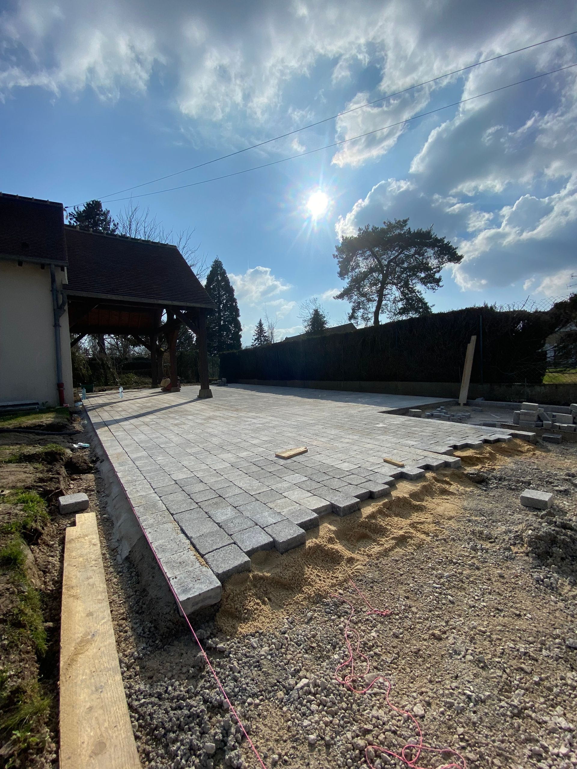 Chantier : terrasse partiellement pavée de dalles grises ; soleil éclatant, ciel bleu.