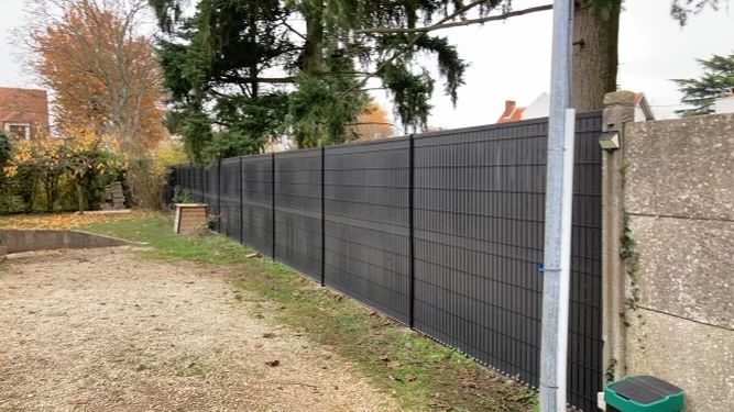 Clôture d'intimité gris foncé longeant une pelouse, avec des arbres et des bâtiments en arrière-plan.
