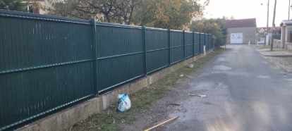 Une clôture métallique verte longe un trottoir à côté d'une route, avec un bâtiment au bout de la route.