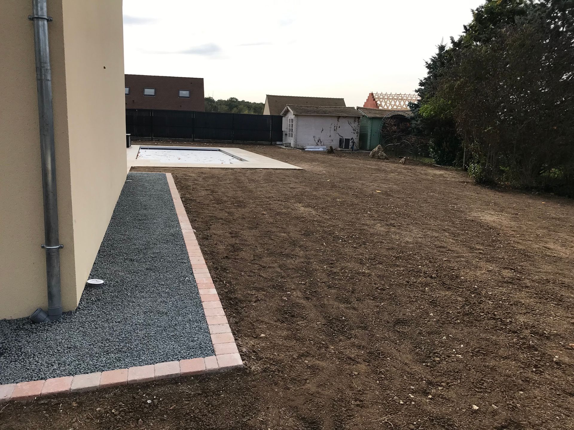 Jardin avec bordure de gravier et pelouse de terre, jouxtant une maison beige. Une piscine et un abri de jardin se devinent au loin.