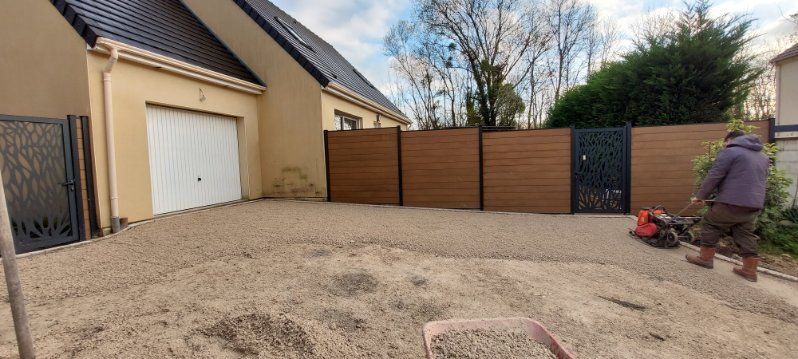 Une personne manipule une plaque vibrante sur du gravier devant une maison.