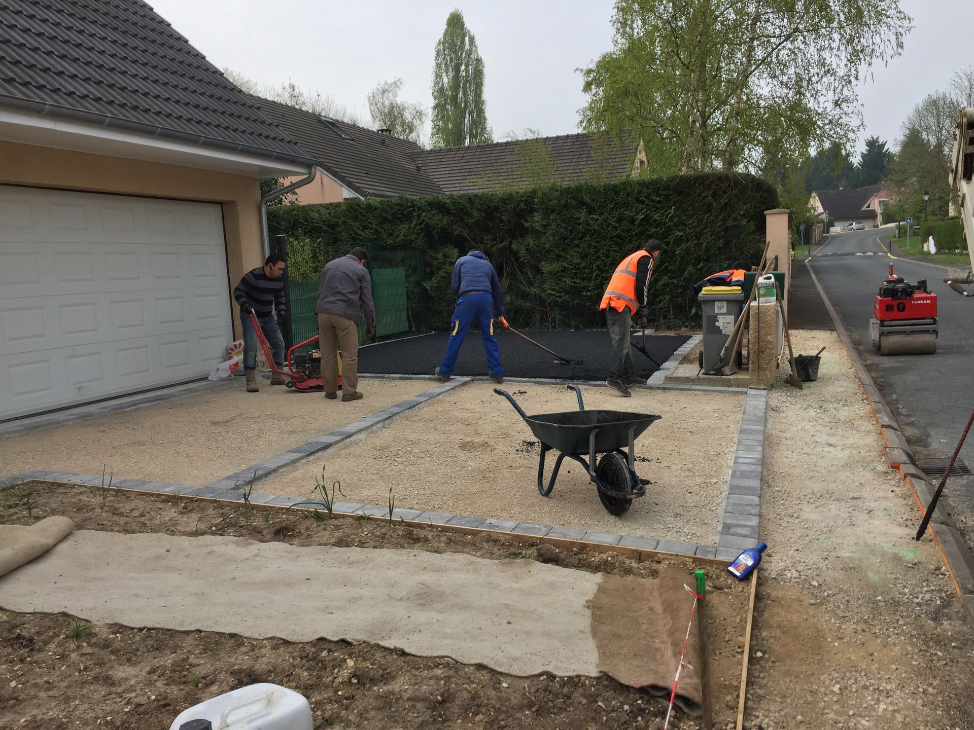 Des ouvriers étalent du gravier pour une allée. Ils utilisent divers outils, dont un compacteur. Scène en extérieur, de jour.