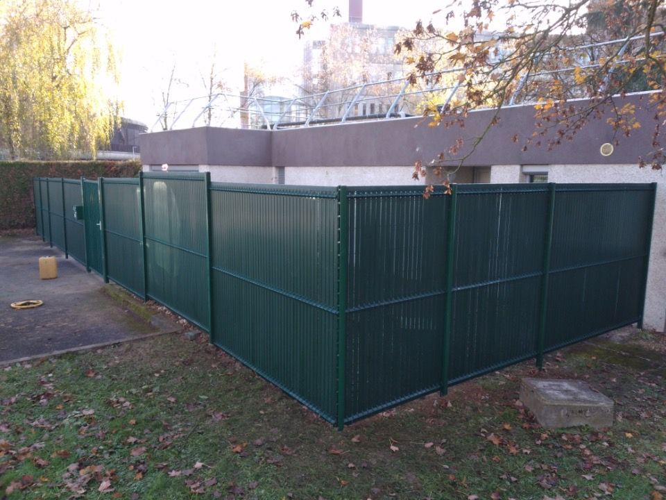 Une clôture à claire-voie vert foncé entoure un espace extérieur jouxtant un bâtiment, sur un terrain herbeux jonché de feuilles mortes.