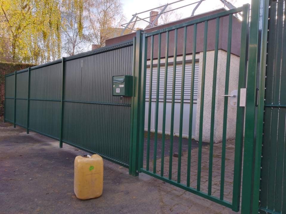 Un portail et une clôture en métal vert précèdent un bâtiment avec une fenêtre fermée et une boîte aux lettres. Un conteneur jaune se trouve à proximité.