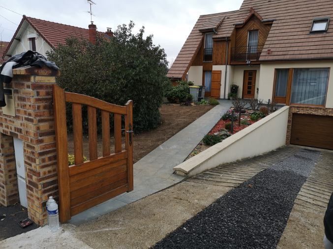 Un portail en bois s'ouvre sur un chemin en béton menant à une maison au toit brun et à une rampe longeant un jardin.