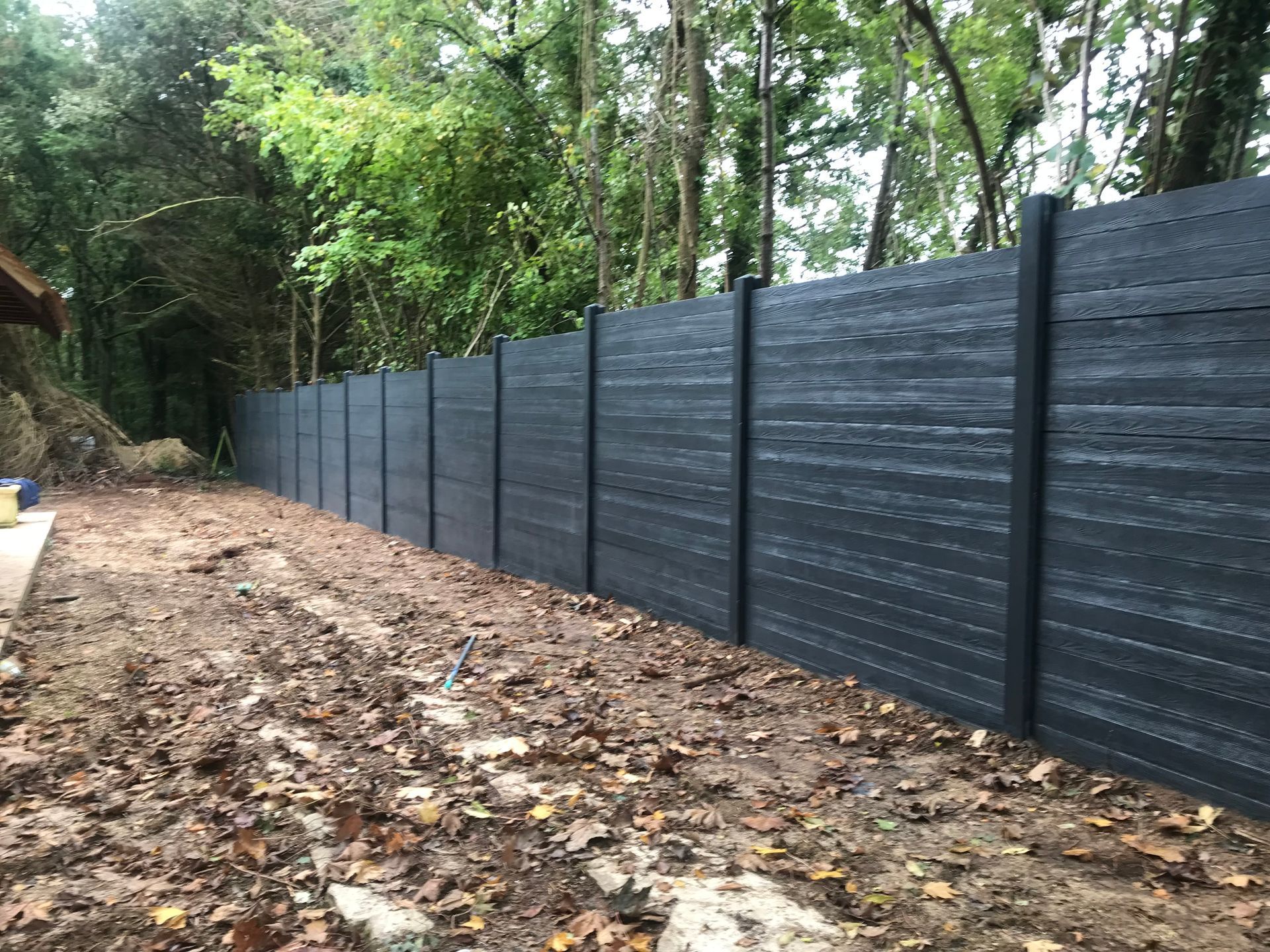 Clôture composite gris foncé dans un jardin boueux, arbres en arrière-plan.