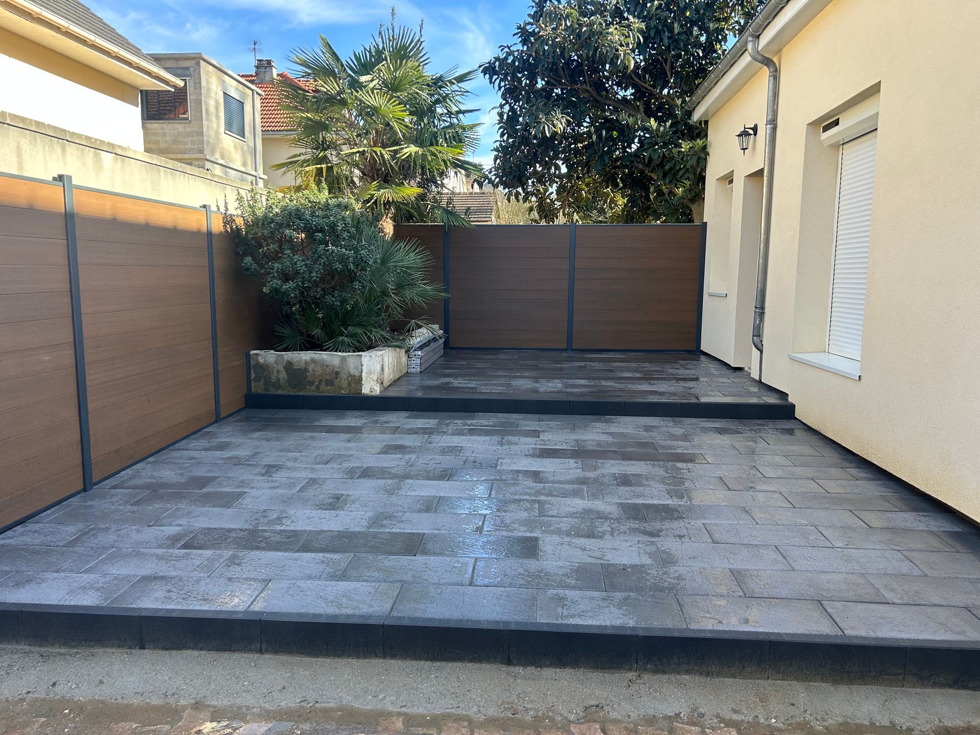 Une terrasse en pierre grise agrémentée d'un petit parterre surélevé, entourée d'une clôture composite marron entre deux bâtiments.
