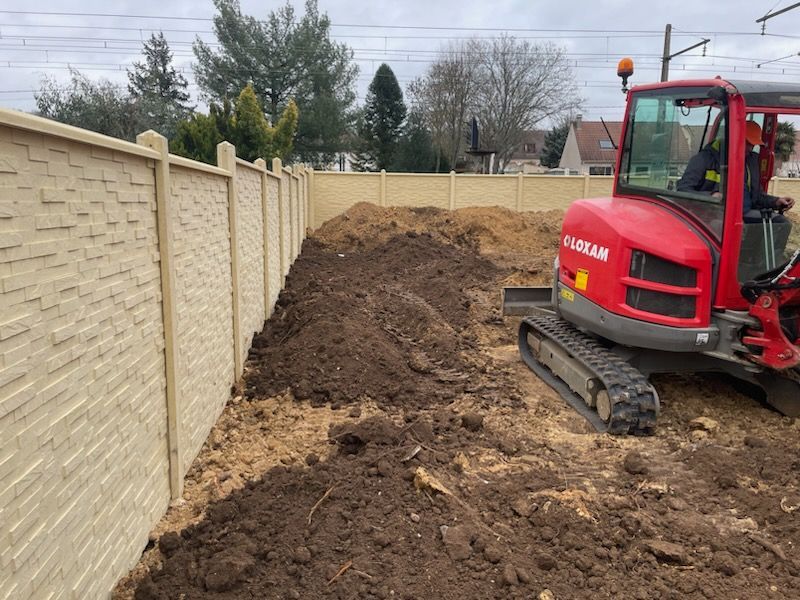 Une mini-pelle rouge creuse dans un jardin entouré d'une clôture en béton. On aperçoit des tas de terre.